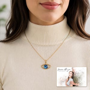 Gold-tone necklace with a blue eye-shaped pendant, worn view. A personalized greeting-card example appears in the corner.