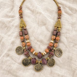 May include: A multi-strand beaded necklace featuring a mix of brown, tan, and purple beads in various shapes and textures. The design includes gold-toned triangular spacers and five circular, textured metal pendants hanging from the lower strands.