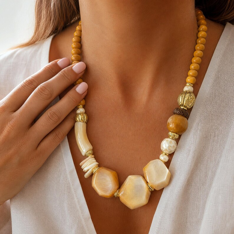 May include: A statement necklace featuring a mix of mustard yellow round beads, textured gold-tone metal spacers, a dark brown porous bead, a smooth cream-coloured cylindrical bead, and three large, irregular, polished cream-coloured geometric pendants.