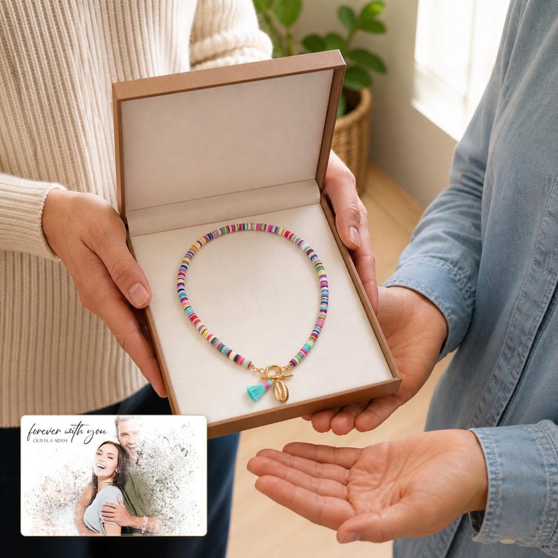 May include: A multi-colored beaded necklace featuring small flat circular beads in various hues, finished with a gold-toned toggle clasp, a small light blue tassel, and a seashell charm, presented inside an open brown gift box with a white interior.