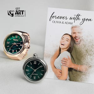 Peut inclure: Deux bagues-montres et une carte avec le texte "forever with you". Une bague-montre est en or rose avec un cadran vert, l'autre est en argent avec un cadran vert. La carte pr&eacute;sente une photo d'un couple.