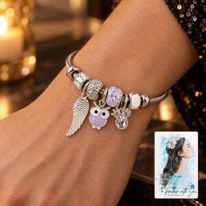 May include: A silver-toned charm bracelet featuring a twisted cable band. The bracelet includes several charms: a silver wing, a purple and white owl, a sparkling crystal bead, a light purple floral bead, and a small crown-shaped crystal charm.