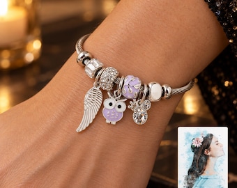 Könnte beinhalten: Ein silberfarbenes Bettelarmband mit einem gedrehten Kabelband. Das Armband enthält mehrere Anhänger: einen silbernen Flügel, eine lila-weiße Eule, eine funkelnde Kristallperle, eine helllila Blumenperle und einen kleinen kronenförmigen Kristallanhänger.