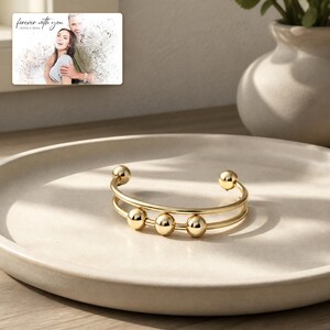 May include: A gold-toned cuff bracelet featuring two parallel bands and three movable spherical beads along the center. In the background, a rectangular card displays a photograph of a couple with the text "forever with you" and "OLIVIA & ADAM".
