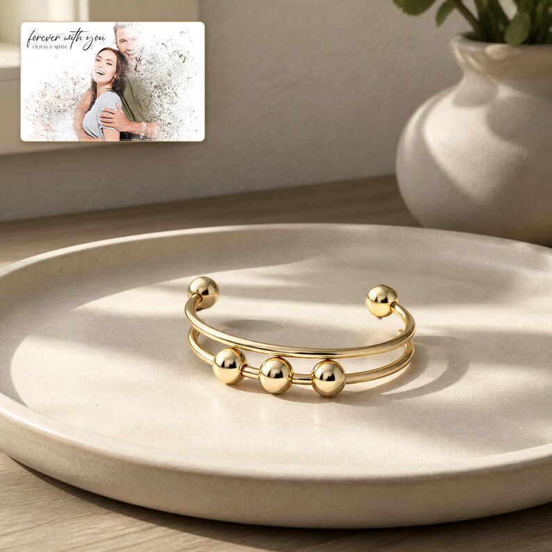 May include: A gold-toned cuff bracelet featuring two parallel bands and three movable spherical beads along the center. In the background, a rectangular card displays a photograph of a couple with the text "forever with you" and "OLIVIA & ADAM".