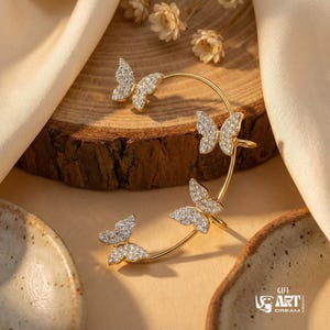 May include: A gold ear cuff with four crystal-encrusted butterfly designs. The butterflies are set along a curved bar, creating an elegant accessory. The cuff is displayed on a wooden surface. The background includes a beige cloth.