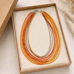 May include: A multi-strand necklace featuring various shades of orange, yellow, and amber beads. The design includes several layers of small, round, and faceted beads, with one thin metallic chain strand, all secured by a silver-toned clasp.