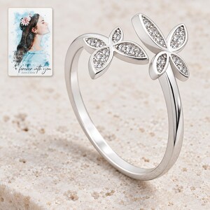 Double butterfly open ring with clear-accent wings displayed on warm ivory stone style. A personalized digital greeting card is shown alongside.