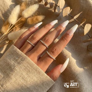 May include: A hand wearing three silver rings with a textured design. The rings are on a hand with long, white, polished nails. The background includes dried flowers and a beige fabric.