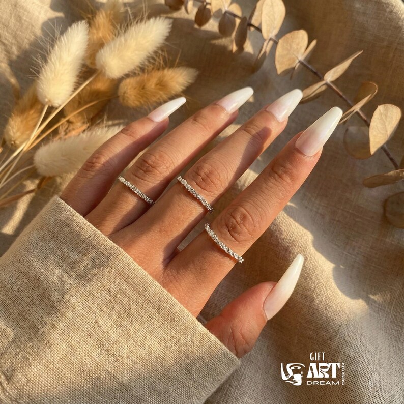 May include: A hand wearing three silver rings with a textured design. The rings are on a hand with long, white, polished nails. The background includes dried flowers and a beige fabric.