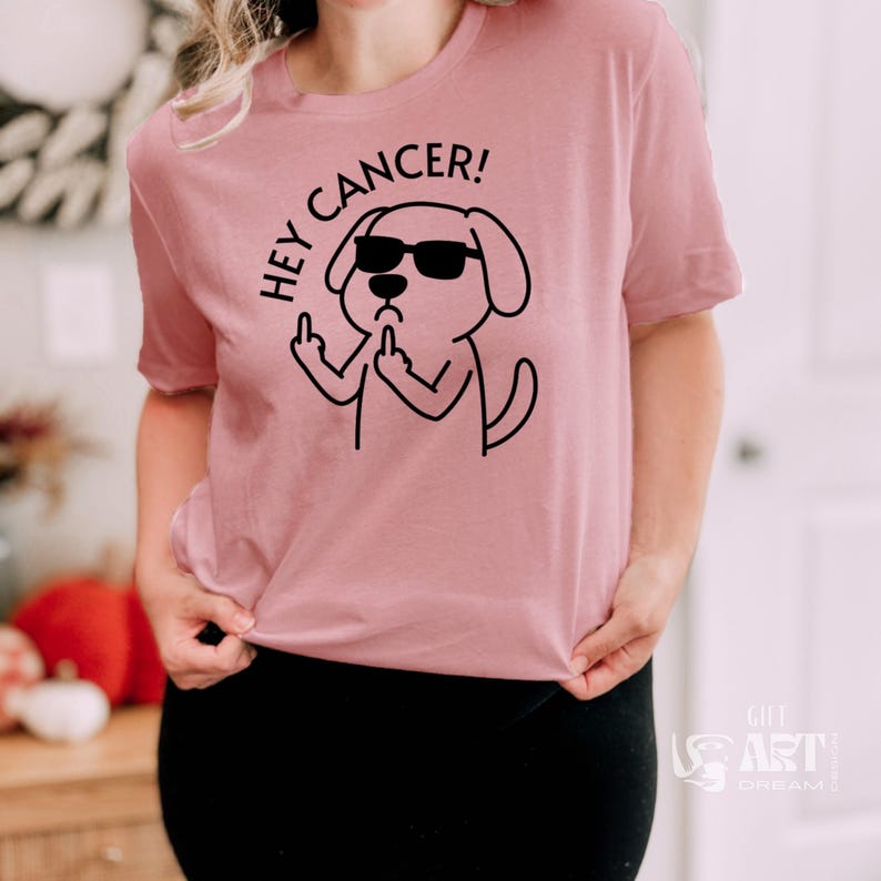 May include: Pink t-shirt with a black cartoon dog wearing sunglasses and giving the middle finger. The text "Hey Cancer!" is above the dog.