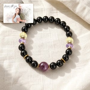 May include: A beaded bracelet featuring smooth black spherical beads, two faceted yellow beads, two faceted purple beads, and one larger faceted dark pink bead. Small metallic gold-toned spacers are placed between the beads. A rectangular card shows a photo of a couple with the text "forever with you" and "OLIVIA & ADAM".