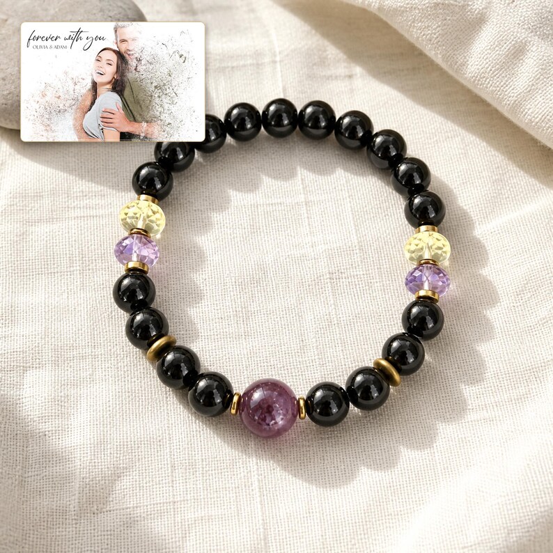 May include: A beaded bracelet featuring smooth black spherical beads, two faceted yellow beads, two faceted purple beads, and one larger faceted dark pink bead. Small metallic gold-toned spacers are placed between the beads. A rectangular card shows a photo of a couple with the text "forever with you" and "OLIVIA & ADAM".