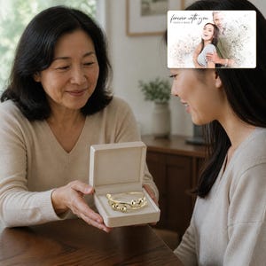 May include: A gold-toned metal bangle bracelet featuring four smooth, round beads along the front, presented inside a square, light beige hinged jewelry box. The bracelet has a polished finish and a simple, open-ended design with rounded ends.