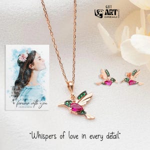 May include: Rose gold hummingbird necklace and earrings set with ruby and emerald accents. The necklace features a pendant with a ruby body and emerald wings. The earrings mirror the pendant design. The image also includes a watercolor portrait and the text "forever with you".