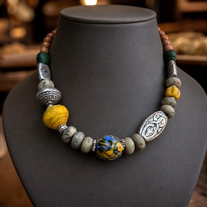 Puede incluir: Un collar de cuentas con una mezcla de cuentas de madera redondas, cuentas de piedra gris lisa y espaciadores de metal plateado texturizado. El dise&ntilde;o incluye una cuenta amarilla grande, una cuenta de vidrio moteada azul y amarilla y detalles plateados.