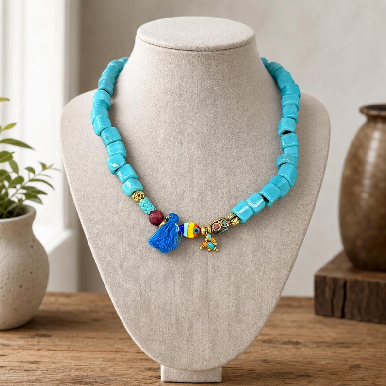 May include: A necklace featuring chunky, square-shaped turquoise-colored beads. The center includes a small blue fabric tassel, a multi-colored glass fish charm, and gold-toned metal spacers with small embedded stones, all arranged on a cord.