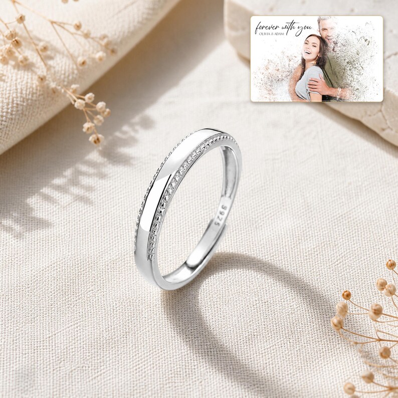 May include: A silver ring with a smooth center band and textured beaded edges, featuring an S925 hallmark stamp. In the upper corner, a rectangular card displays a photograph of a couple with the text "forever with you" and "OLIVIA & ADAM".