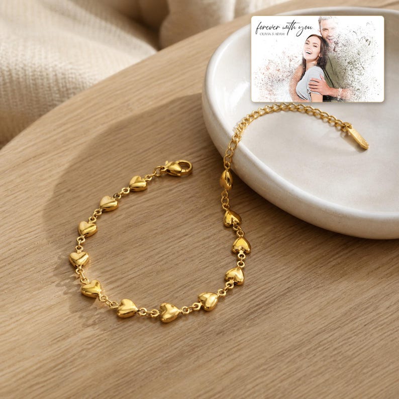 May include: A gold-toned chain bracelet featuring a series of small, polished heart-shaped links. The bracelet is displayed alongside a rectangular card showing a photograph of a couple with the text "forever with you" and "OLIVIA & ADAM".