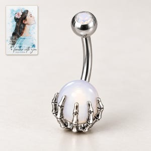 White-ball skeleton-claw barbell on an ivory background, with its small faceted top stone style visible. A personalized digital greeting card is shown alongside.