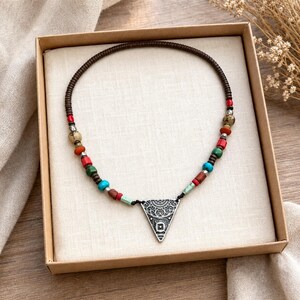 Op de afbeelding: Een ketting met een donkerbruin koord geregen met kleine, veelkleurige kralen in tinten rood, turkoois, groen en bruin, geaccentueerd door zilverkleurige tussenstukjes en een centrale zilverkleurige driehoekige hanger met patroon.
