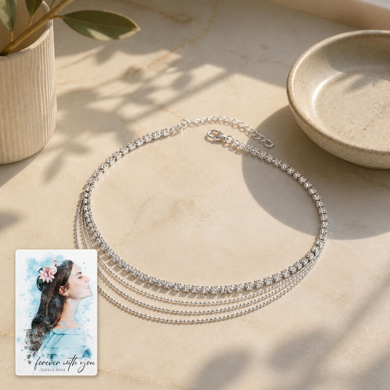 May include: A silver-toned multi-strand necklace featuring one row of clear round rhinestones and three delicate beaded chains. Beside the necklace is a rectangular card displaying a watercolour illustration of a person and the text "forever with you".