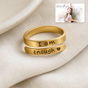 K&ouml;nnte beinhalten: Ein goldfarbener Wickelring mit der Gravur "I am enough" und einem kleinen Herzsymbol. Der Ring hat eine glatte, polierte Oberfl&auml;che und ein offenes Design. Eine kleine rechteckige Karte mit einem Paarfoto und dem Text "forever with you" ist in der oberen rechten Ecke zu sehen.