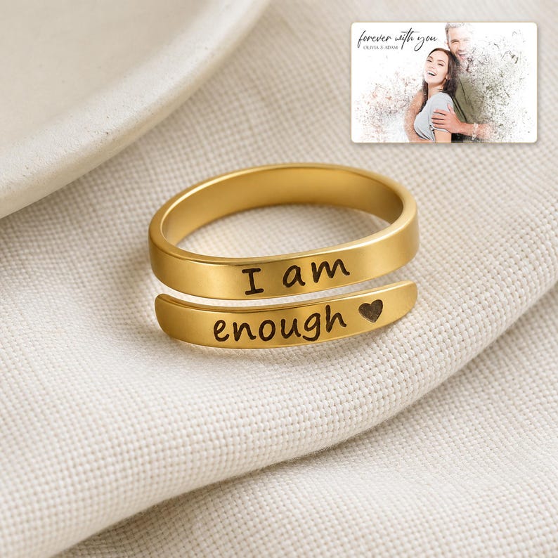 K&ouml;nnte beinhalten: Ein goldfarbener Wickelring mit der Gravur "I am enough" und einem kleinen Herzsymbol. Der Ring hat eine glatte, polierte Oberfl&auml;che und ein offenes Design. Eine kleine rechteckige Karte mit einem Paarfoto und dem Text "forever with you" ist in der oberen rechten Ecke zu sehen.