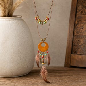 May include: A multi-strand necklace featuring a brown cord, a small crescent-shaped accent with red, green, and gold-toned beads, and a large circular wooden pendant adorned with dangling red and turquoise beads, metal charms, and brown feathers.