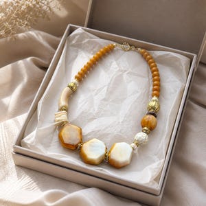 May include: A beaded necklace featuring a mix of amber-colored round beads, smooth cream-colored hexagonal stones, and textured gold-toned metal spacers. The necklace is arranged in a circular shape inside a square gift box lined with white tissue paper.