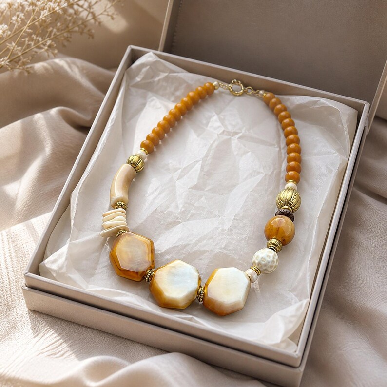 May include: A beaded necklace featuring a mix of amber-colored round beads, smooth cream-colored hexagonal stones, and textured gold-toned metal spacers. The necklace is arranged in a circular shape inside a square gift box lined with white tissue paper.