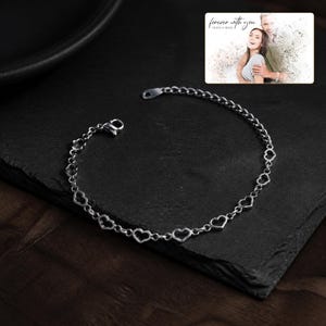 May include: A silver-toned chain bracelet featuring a series of interconnected open heart-shaped links. In the upper right corner, a rectangular card displays a black and white photograph of a couple with the text "forever with you" and "OLIVIA & ADAM".