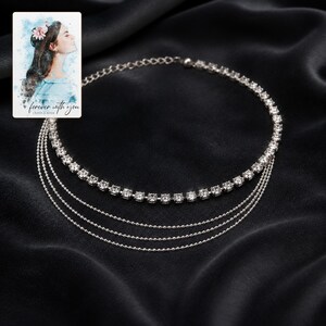 May include: A silver-toned multi-strand necklace featuring one row of clear square-cut rhinestones and three delicate beaded chains. Included is a small rectangular card with a watercolour illustration of a person and the text forever with you.