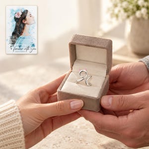 Two adults exchange an open taupe styling box containing one silver-tone cat-curl ring. A personalized digital greeting card is shown alongside.