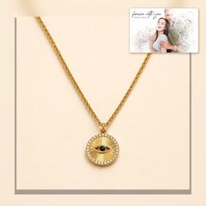 Gold-tone necklace with a round sunburst and black eye detail, framed product view. A personalized greeting-card example appears in the corner.
