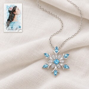 Winter Azure Snowflake Necklace arranged on soft ivory linen in a refined neutral flat lay. A personalized digital greeting card is shown alongside.