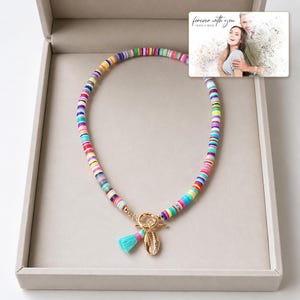 May include: A necklace made of multicolored flat circular beads featuring a gold-toned toggle clasp, a small turquoise tassel, and a gold-toned cowrie shell charm. A rectangular card displays a photo of a couple with the text "forever with you".