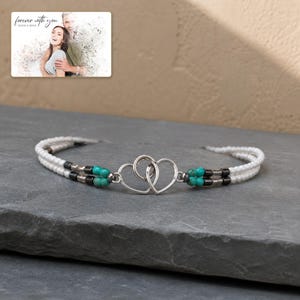 May include: A double-strand beaded bracelet featuring two interlocking silver-toned heart charms at the centre. The bracelet is composed of white seed beads, metallic grey cylindrical beads, and bright turquoise round beads. A photo card reads "forever with you".