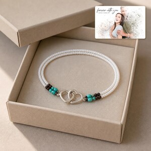 May include: A double-strand bracelet featuring small white beads, accented with turquoise and dark brown beads near a silver-toned interlocking heart clasp. The bracelet is presented in a box with a card showing a photo and the text "forever with you".