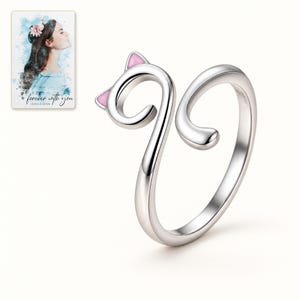 Single open cat-curl ring on an ivory-white background, showing the two soft pink ears and curved tail. A personalized digital greeting card is shown alongside.