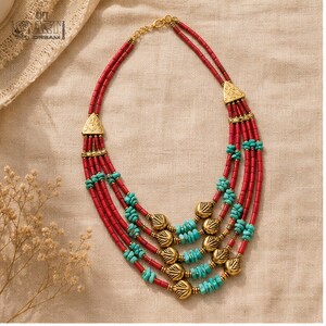 May include: A multi-strand necklace featuring cylindrical red beads and small turquoise-colored stone chips. The design includes gold-toned metal spacers and triangular connectors, with gold-toned leaf-shaped charms integrated into the lower strands.