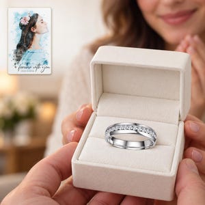 An adult receiving an open ivory ring box with the silver-tone clear-accent band prominent in the foreground. A personalized digital greeting card is shown alongside.