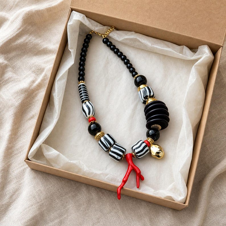 Op de afbeelding: Een ketting met zwarte ronde kralen, zwart-wit gestreepte cilindrische kralen, een stapel zwarte schijfkralen, kleine rode accentkralen en een felrode takvormige hanger, verbonden door goudkleurige metalen tussenstukjes en sluitingen.