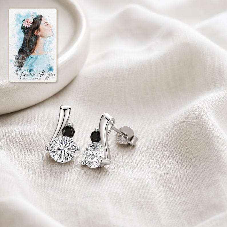 K&ouml;nnte beinhalten: Ein Paar Ohrstecker aus Silber mit einem gro&szlig;en runden klaren Stein und einem kleineren runden schwarzen Stein in einer geschwungenen Metallfassung. Eine rechteckige Karte zeigt eine Aquarellillustration einer Person und den Text forever with you.