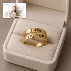 K&ouml;nnte beinhalten: Ein goldfarbener Ring mit zwei B&auml;ndern, graviert mit dem Text "I am" auf dem oberen Band und "enough" gefolgt von einem kleinen Herzsymbol auf dem unteren Band, pr&auml;sentiert in einer beigen, mit Samt ausgekleideten Schmuckschatulle mit Fotokarte.