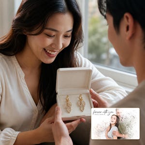 May include: A pair of gold-toned leaf-shaped earrings with embedded clear stones, presented in a cream-colored rectangular jewelry box. Beside the box is a rectangular card featuring a printed photograph of a couple with the text "forever with you".