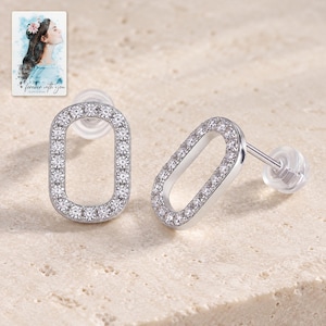 May include: A pair of silver-toned stud earrings featuring an oval-shaped frame encrusted with small clear stones. Each earring includes a post back with a clear silicone clutch. A small card with a watercolor illustration and the text "forever with you" is visible in the upper left corner.