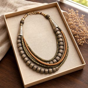Pu&ograve; includere: Una collana a pi&ugrave; fili con perline in legno marrone scuro, perline cilindriche pi&ugrave; piccole color verde oliva e perline rotonde color marrone chiaro. I fili sono collegati da due terminali conici in metallo dorato con motivi incisi.