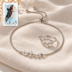 Silver-tone bracelet and open-vine ring styled on textured cream linen, with clear leaf accents facing the camera. A personalized digital greeting card is shown alongside.