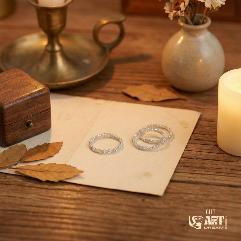 May include: Three silver rings with clear gemstones are displayed on a cream-colored paper. A small wooden box, a brass candle holder, a white candle, and a small vase with dried flowers are also visible on a wooden surface.